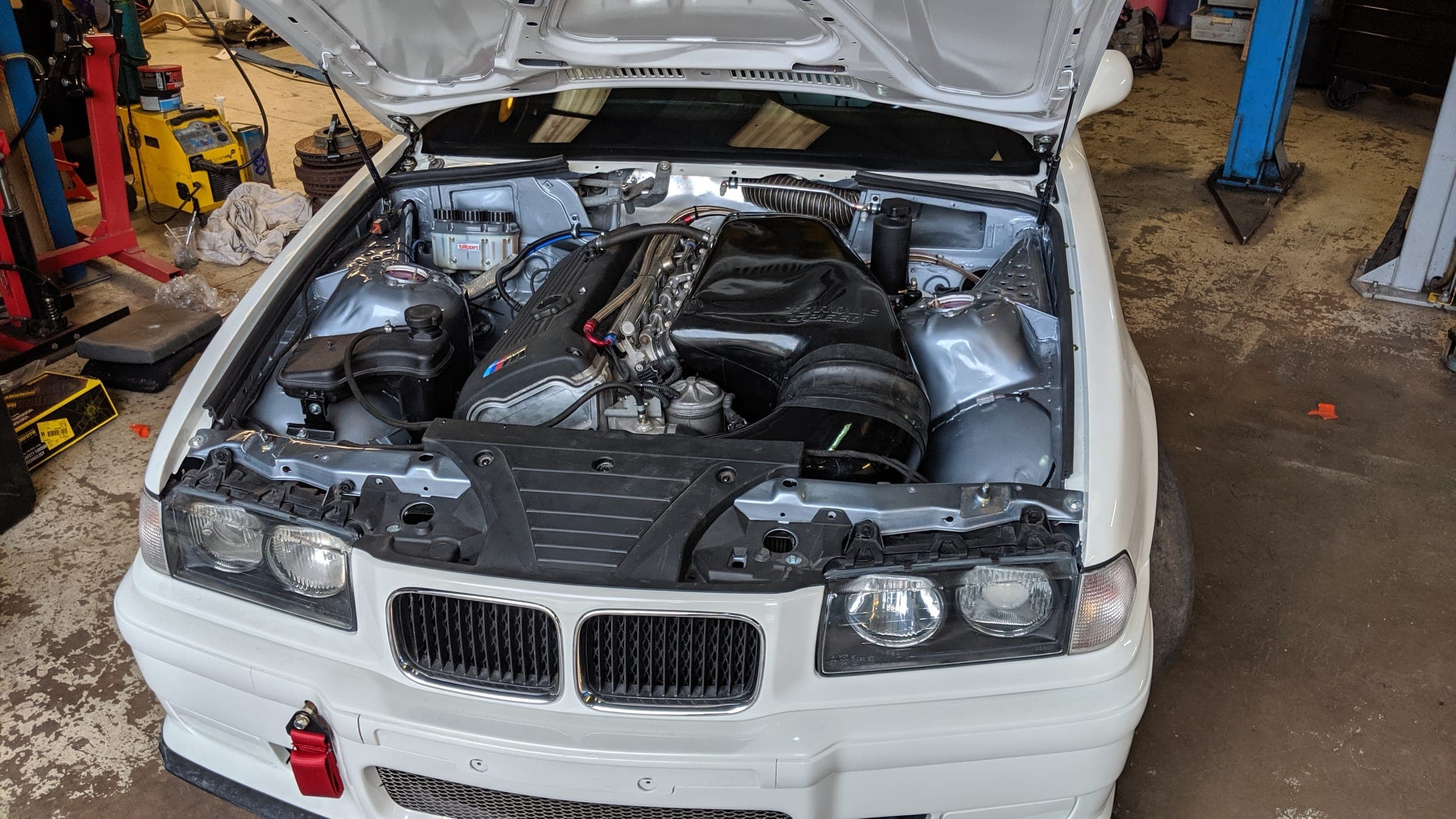 A white BMW E36 with the bonnet up in the workshop